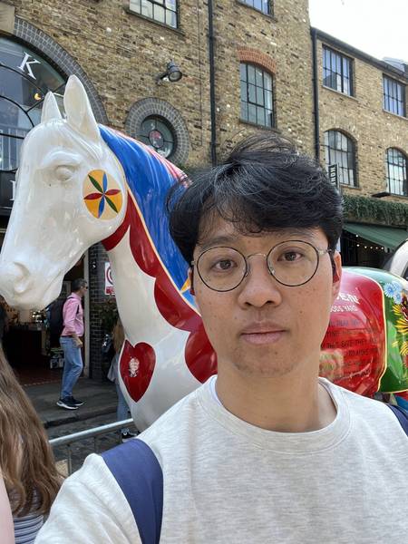 캠든 마켓에서 셀카. 안경 쓴 남성이 흰 티셔츠 입고 카메라를 보고 있다. 뒤로 벽돌 건물과 컬러풀한 조형물이 보인다