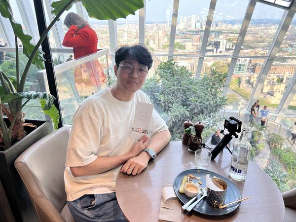 스카이 가든 내부 레스토랑에서 식사 중인 모습. 테이블 위에 음식과 음료가 있고, 뒤로 초록 식물들과 유리창 너머 런던 시내가 보인다