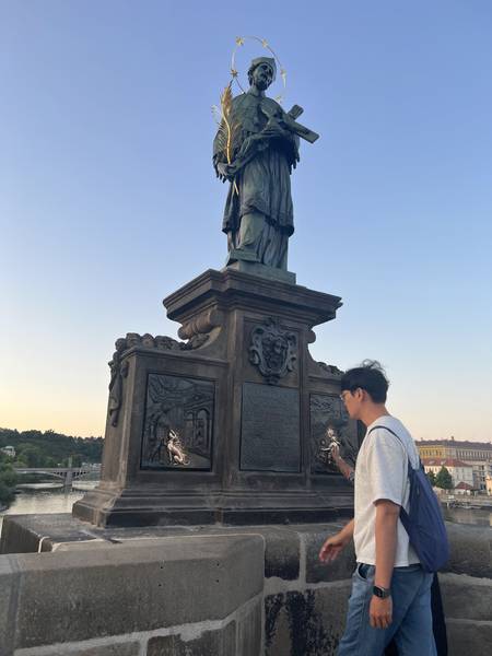 카를교(Charles Bridge) 위 성인 조각상 옆에서 포즈. 석양 빛을 받은 청동 조각상이 실루엣으로 보인다. 하늘이 파란색과 주황색의 그라데이션
