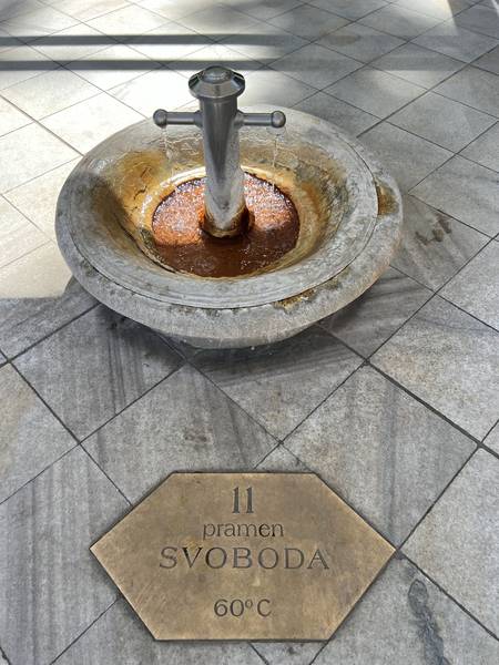 온천수 샘 'Pramen SVOBODA' (자유의 샘, 60°C). 돌로 된 원형 분수에서 뜨거운 물이 나오고, 바닥에 금속 표지판이 박혀 있다