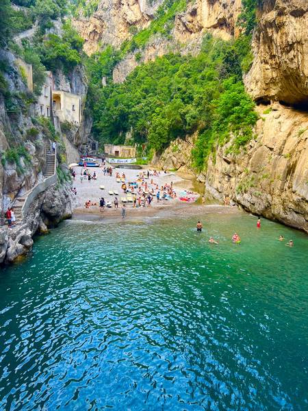 피오르도 디 푸로레(Fiordo di Furore) 해변. 양쪽 절벽 사이 좁은 협곡에 에메랄드빛 바닷물이 차 있다. 절벽에는 계단이 깎여 있고, 작은 자갈 해변에 사람들이 수영하고 있다. 투명한 터키색 물